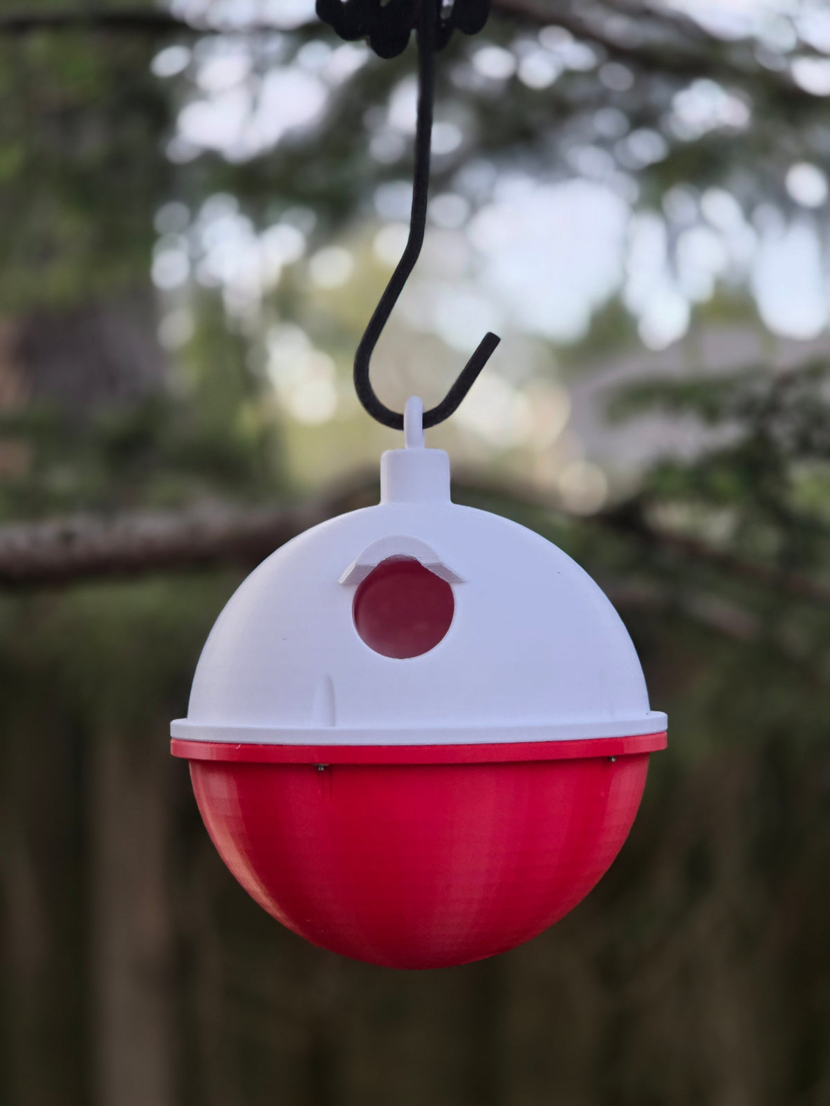 Front view of the Fishing Bobber Bird House on a black hook, round entrance hole centered, blurred yard behind