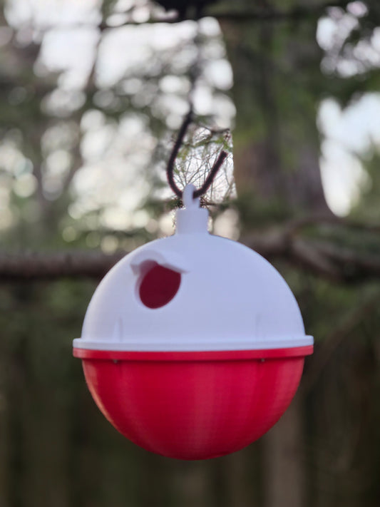 Red-and-white Fishing Bobber Bird House hanging from a hook, entrance hole visible, soft-focus trees behind